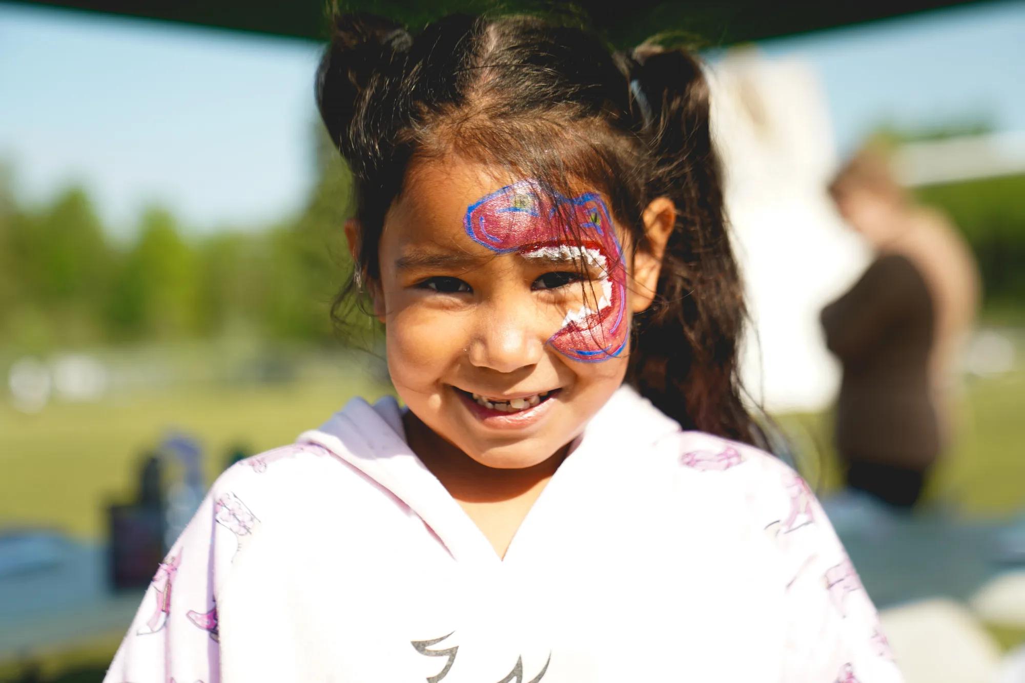Child standing outdoors with colorful face paint on one side of the face, photographed in a park or community event setting with a blurred background.
