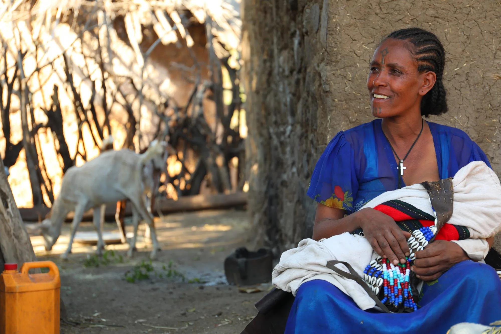 A woman holding a young child in her hands with a goat in the background.