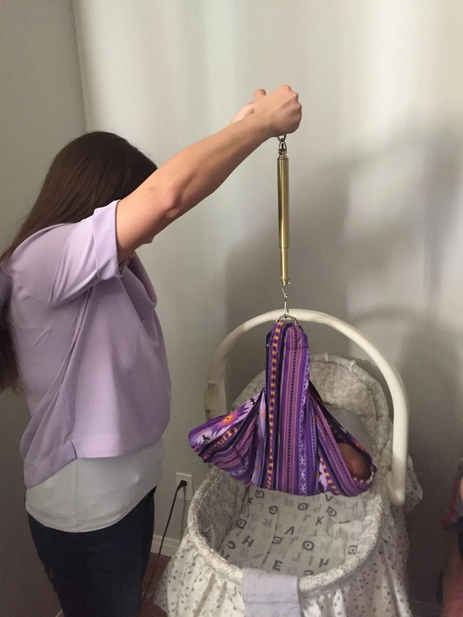 An adult uses a hanging spring scale to weigh an infant secured in a patterned fabric sling suspended above a bassinette indoors.