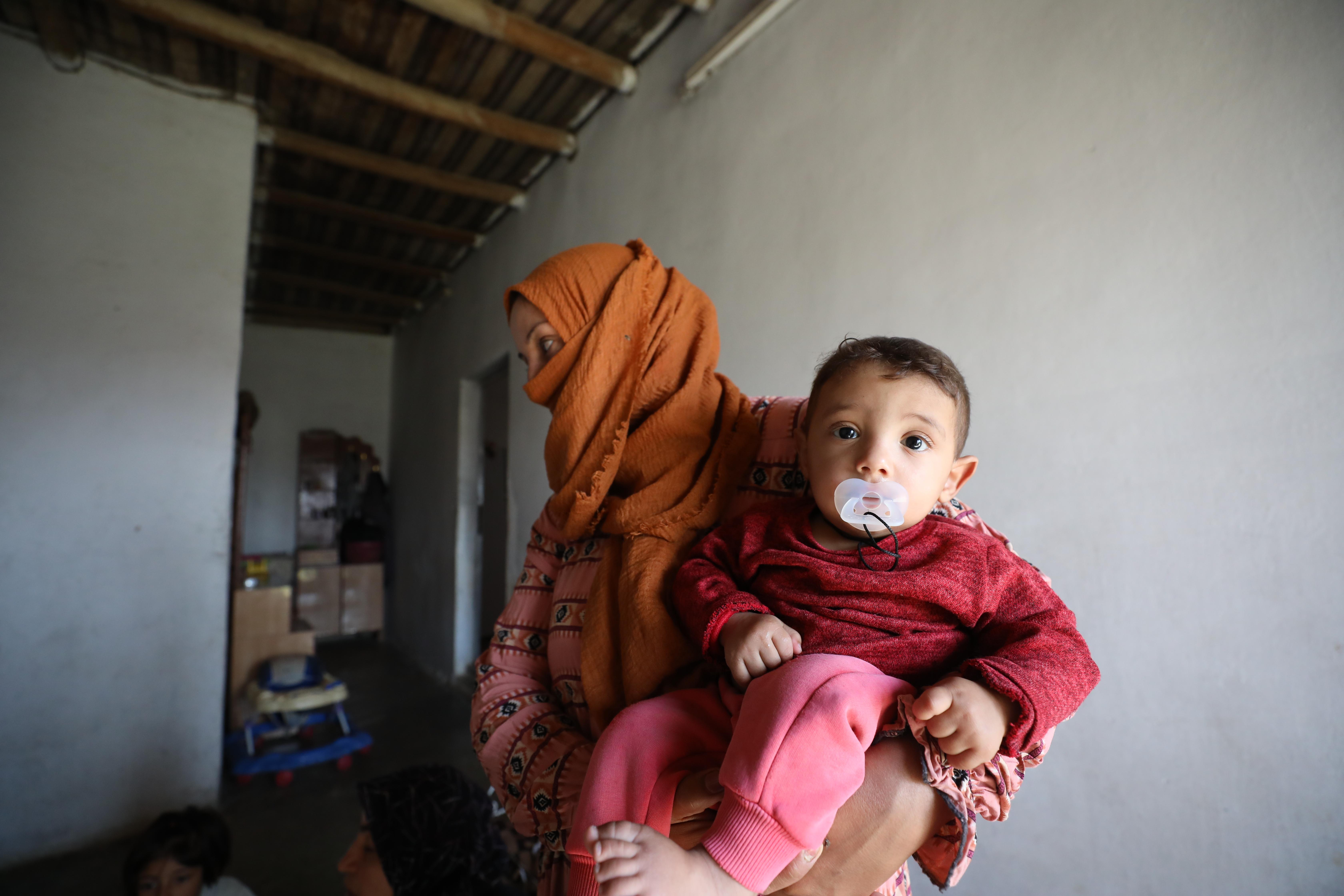 A woman in an orange head scarf holds an infant sucking on a pacifier in her arms.