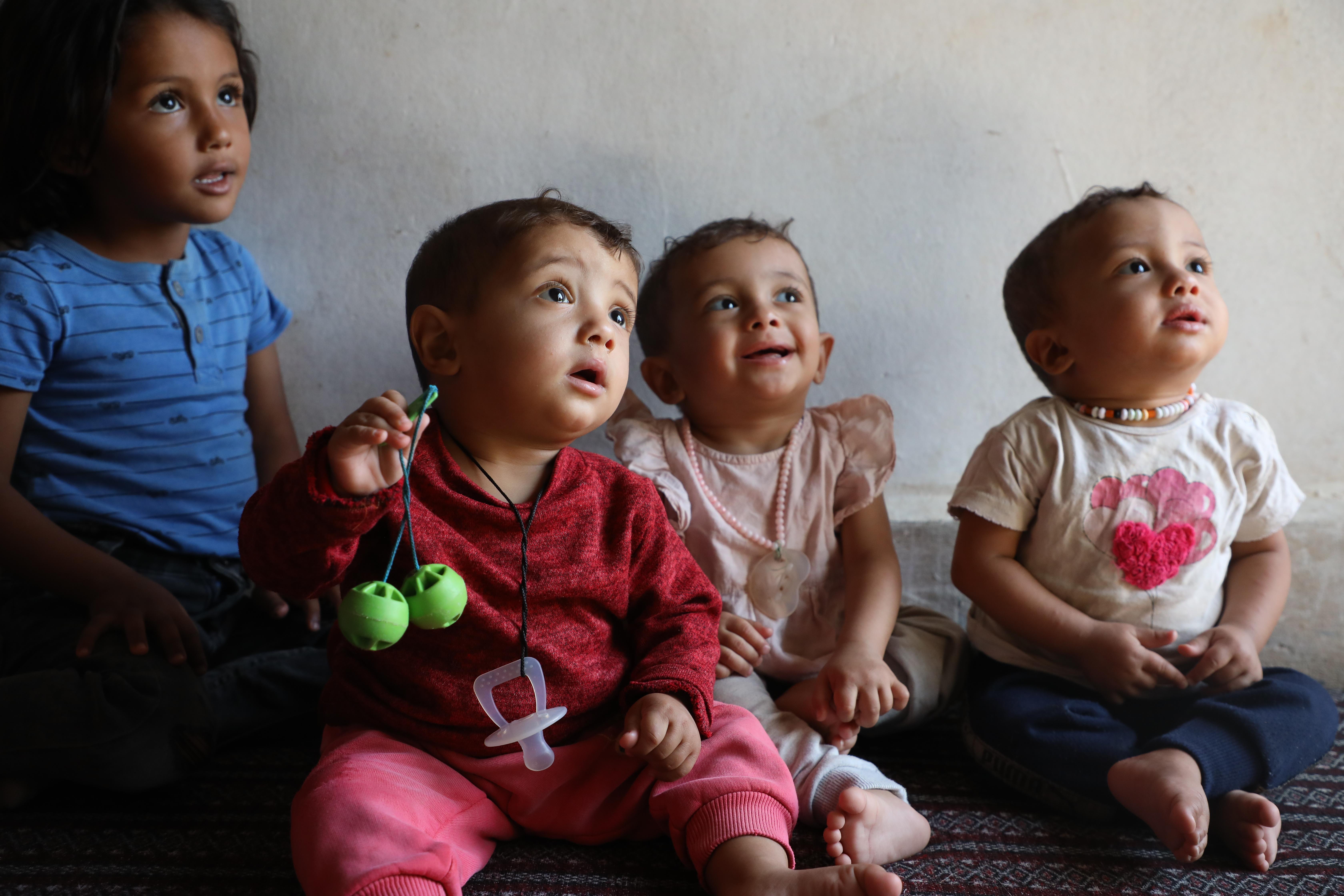 Three small babies, one in red, one in pink, and one in white sit on a mat with an older girl child in blue behind them.