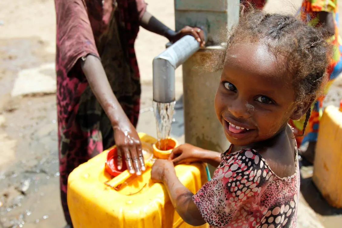 Child standing beside a yellow water container at a community water pump while water flows into a small cup, with additional people and containers visible nearby.