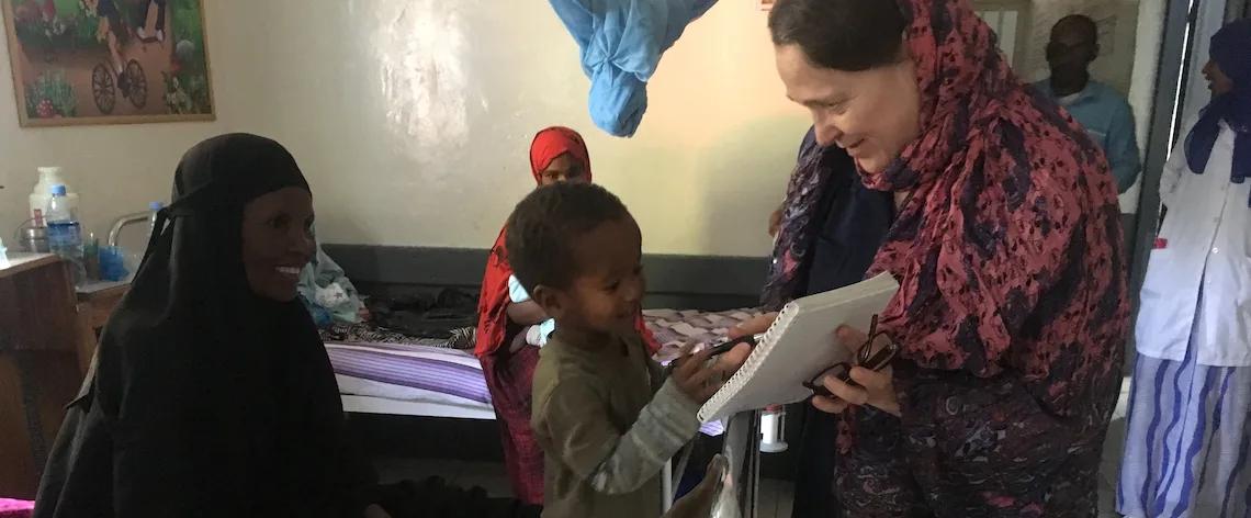 A child stands beside an adult holding a notebook inside a healthcare or community facility, while other people sit and stand nearby around beds and medical equipment.