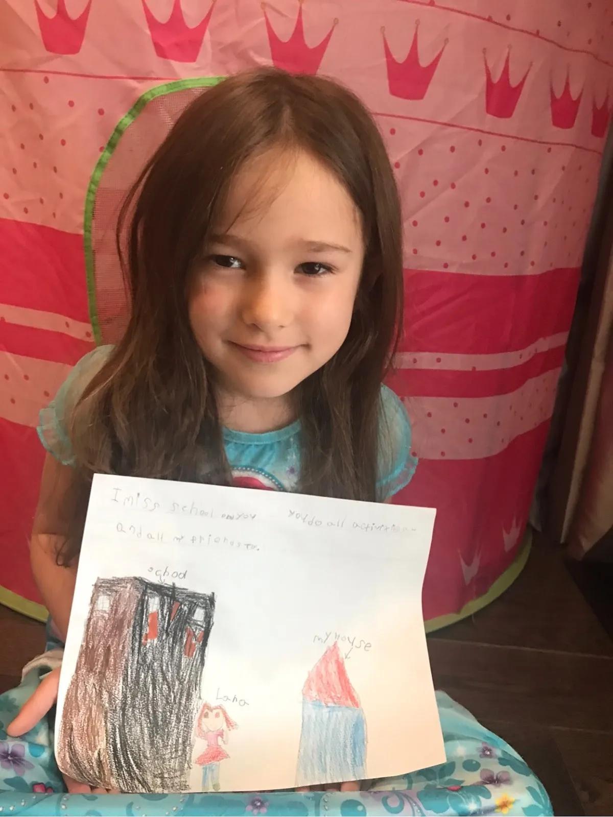 Child seated indoors holding a hand-drawn picture showing a school, a house, and a figure, with handwritten notes on the paper and a pink play tent in the background.