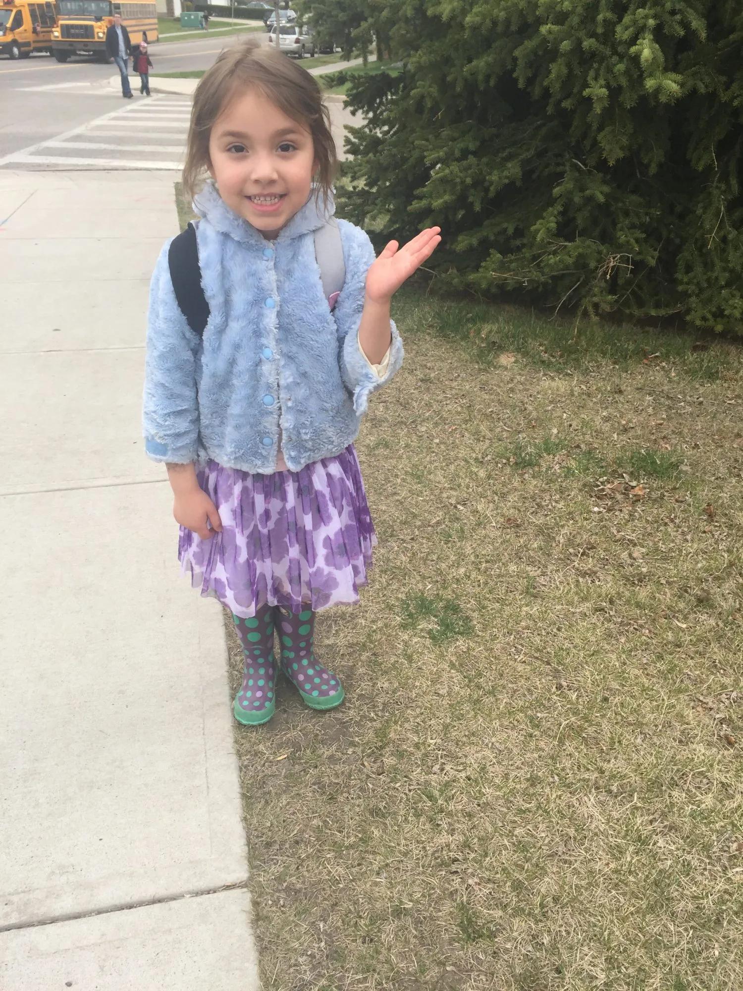 A child wearing a light blue jacket, patterned skirt, and rain boots stands on a sidewalk near a school crosswalk and yellow school buses, with a backpack visible over the shoulders.