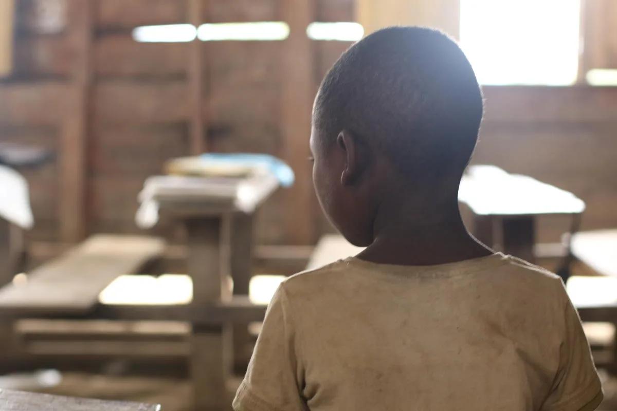 Child viewed from behind standing inside a classroom with wooden desks and benches, illuminated by sunlight entering through windows in the background.