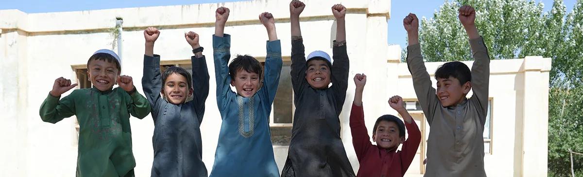 Group of children standing outdoors in front of a building with their arms raised above their heads.
