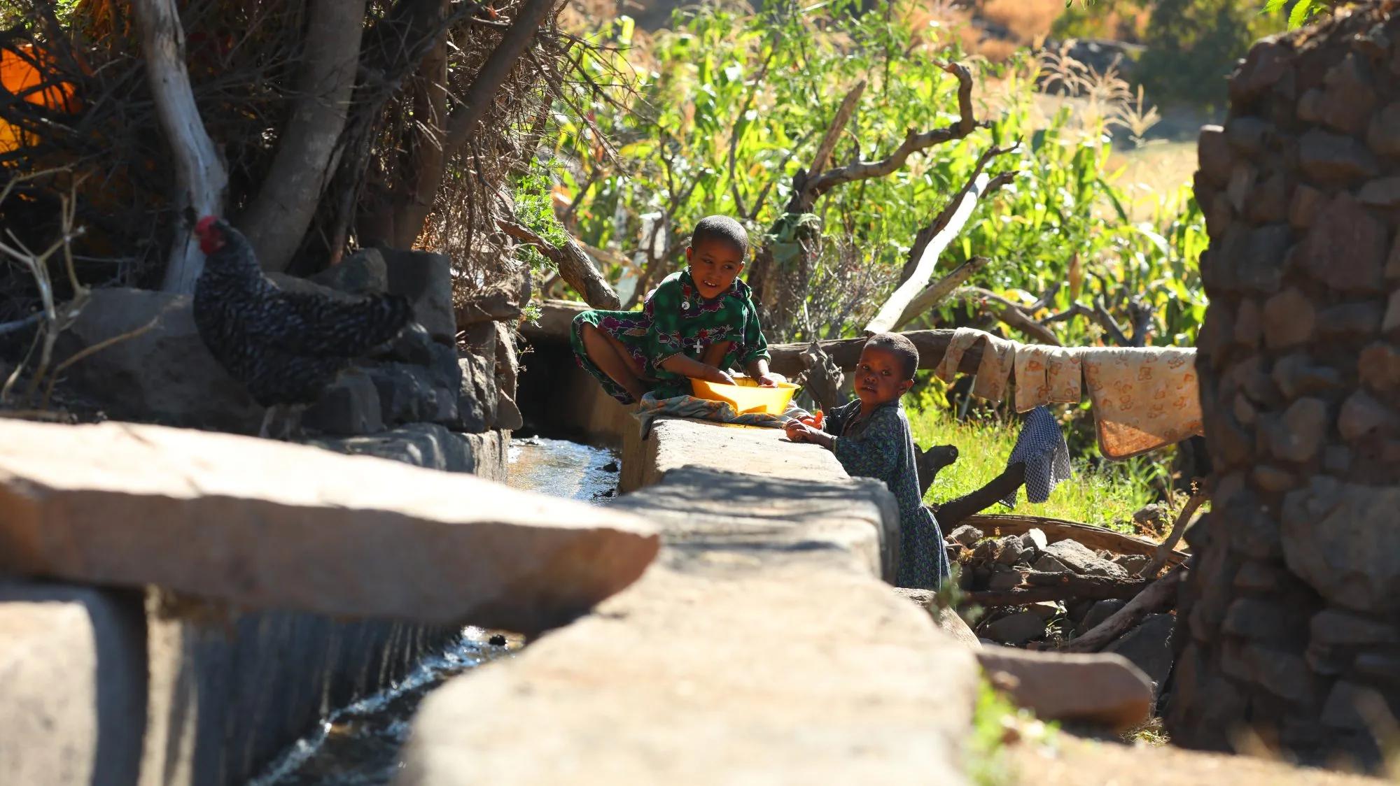 Two children beside a flowing water channel in a rural setting, using a yellow container near vegetation and stone structures.