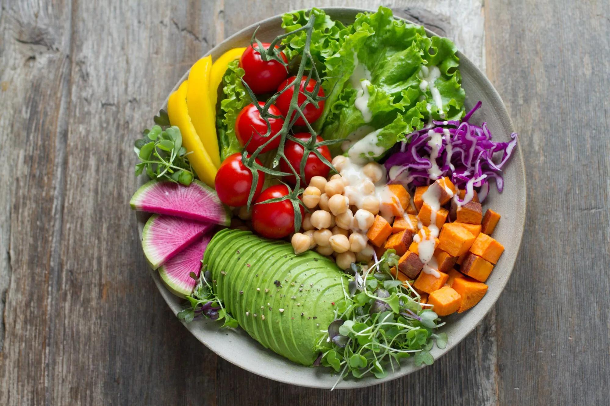 Overhead view of a vibrant plant-based meal bowl containing an arrangement of vegetables and a creamy dressing on a wooden table.