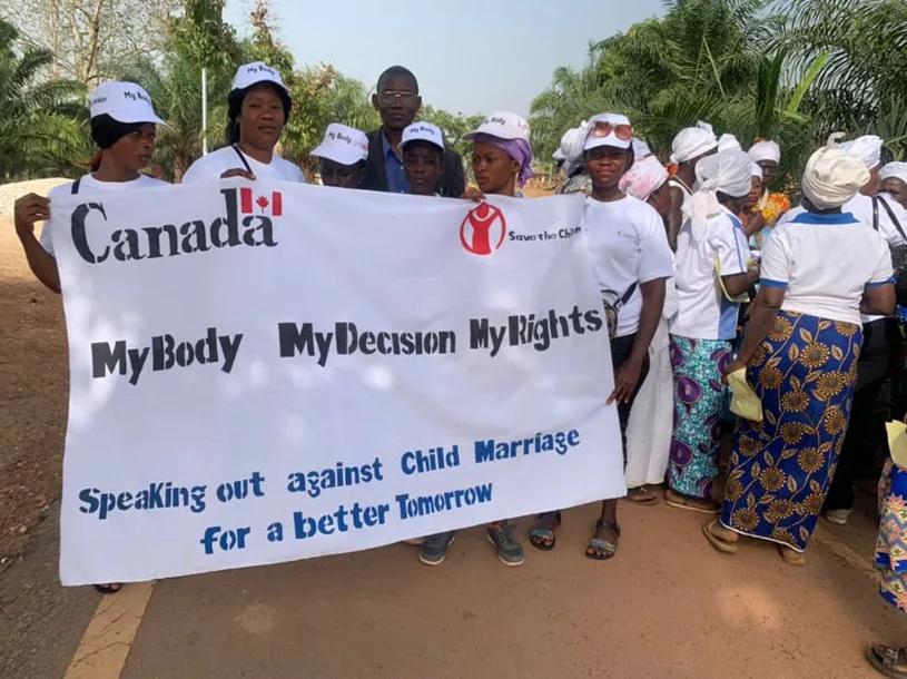 Community members standing outdoors holding a banner during an awareness event. The banner includes the messages ���My Body My Decision My Rights��� and ���Speaking out against Child Marriage for a better Tomorrow.���