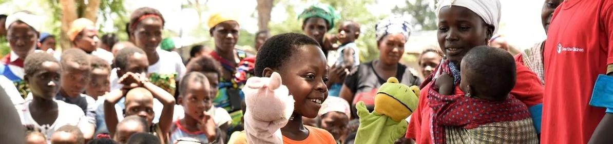 Child holding hand puppets during a community activity, surrounded by children and adults gathered outdoors in a group setting.