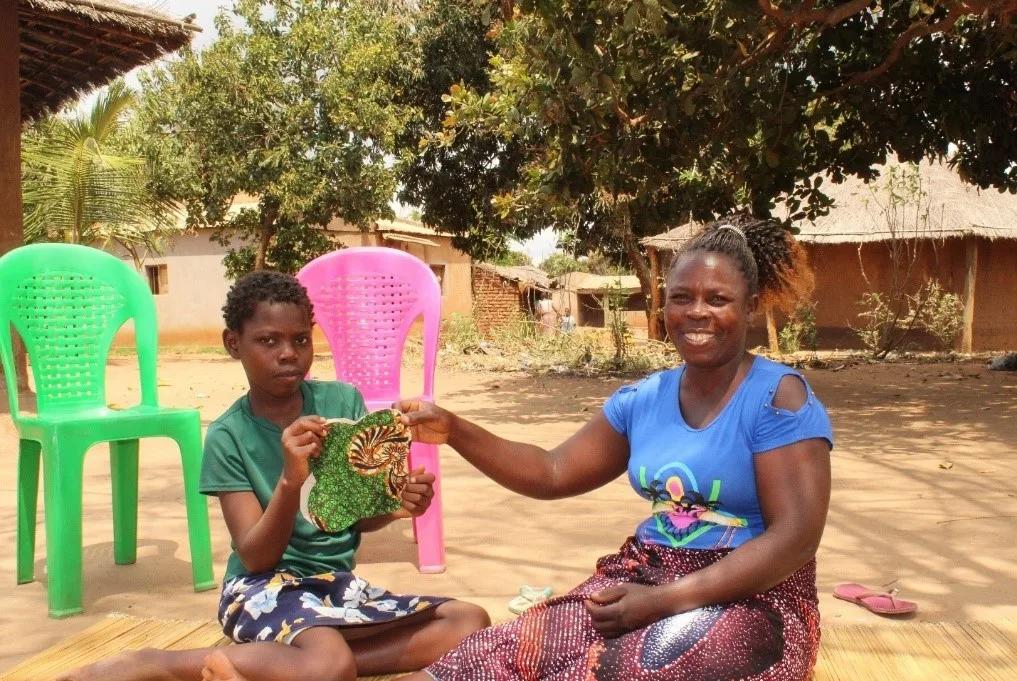 Two individuals seated outdoors in a village setting holding a piece of patterned fabric, with colorful plastic chairs and homes visible in the background.