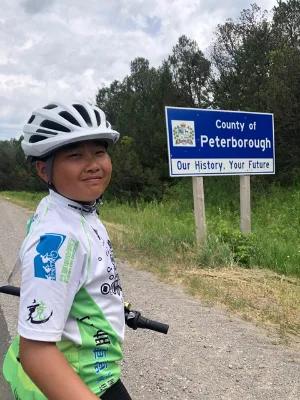 Person wearing a bicycle helmet and cycling jersey standing beside a roadside sign that reads ���County of Peterborough ��� Our History, Your Future,��� with trees and grassy landscape in the background.