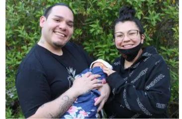 Two adults standing outdoors in front of greenery while holding an infant wrapped in a blanket, posing together for a photograph.