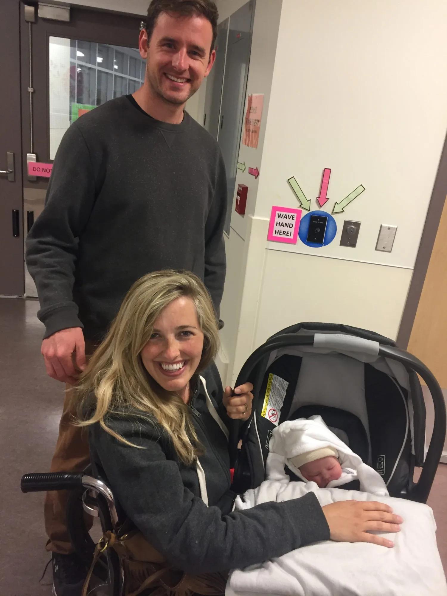 Adult pushing a wheelchair beside an infant car seat containing a newborn wrapped in a blanket, photographed in a hospital corridor during discharge from maternity care.