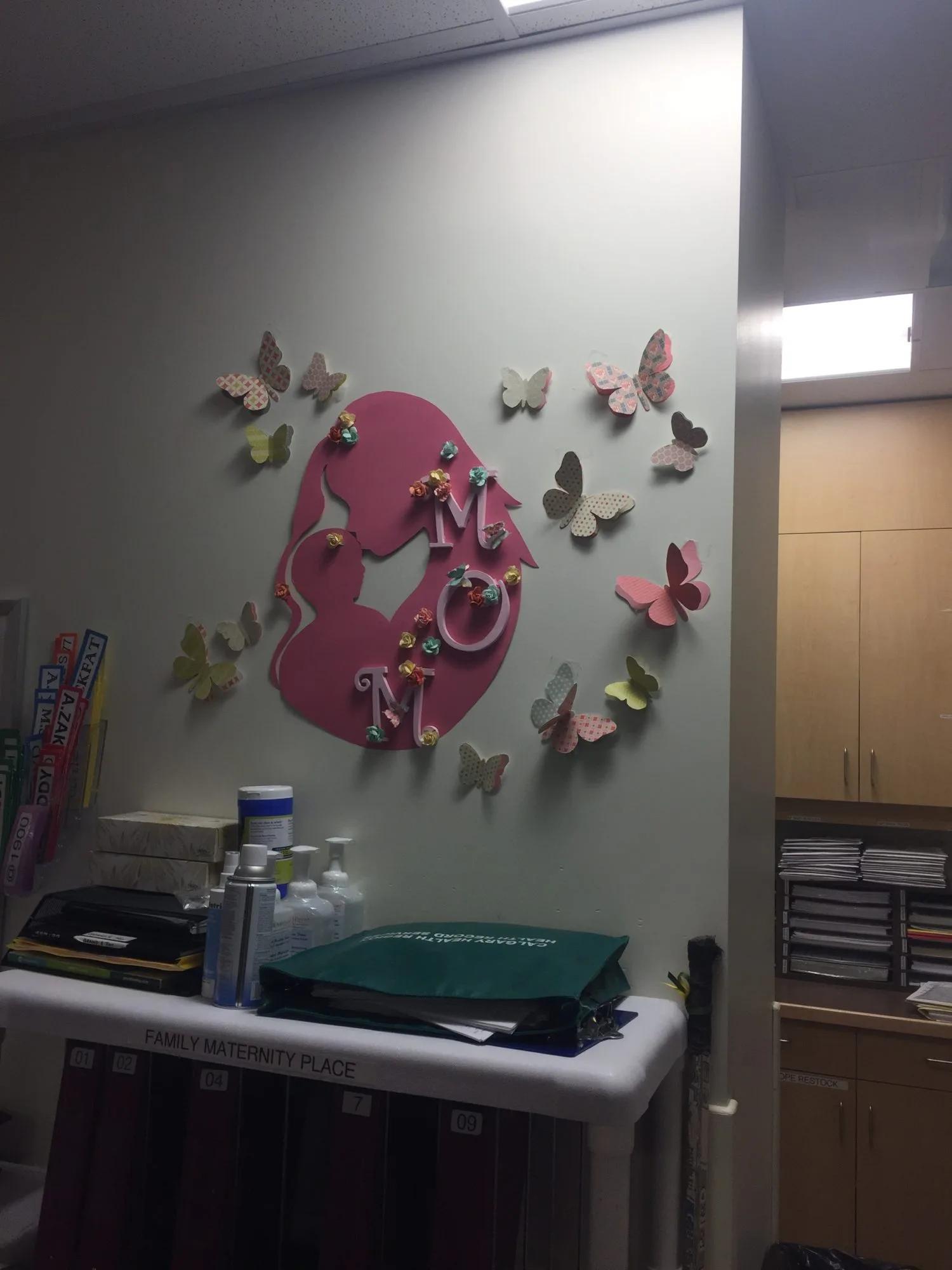 A maternity care area featuring a decorative wall display with a parent-and-infant silhouette, paper butterflies, and healthcare supplies organized on a workstation.