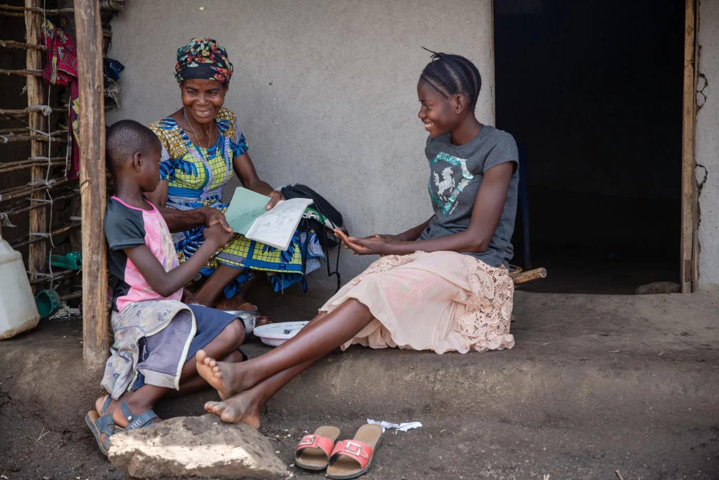 Three people sit together outside a home reviewing papers and a notebook, with one person holding educational materials while the others look on.