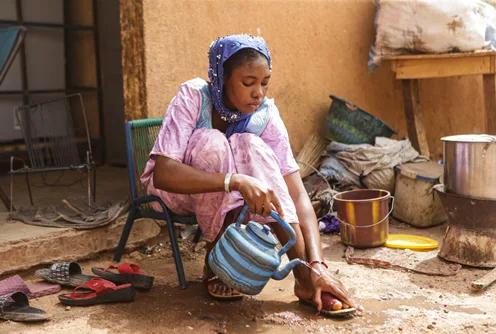 Individual seated outdoors in a courtyard pouring water from a kettle onto a foot, with cooking pots, containers, and household items nearby.