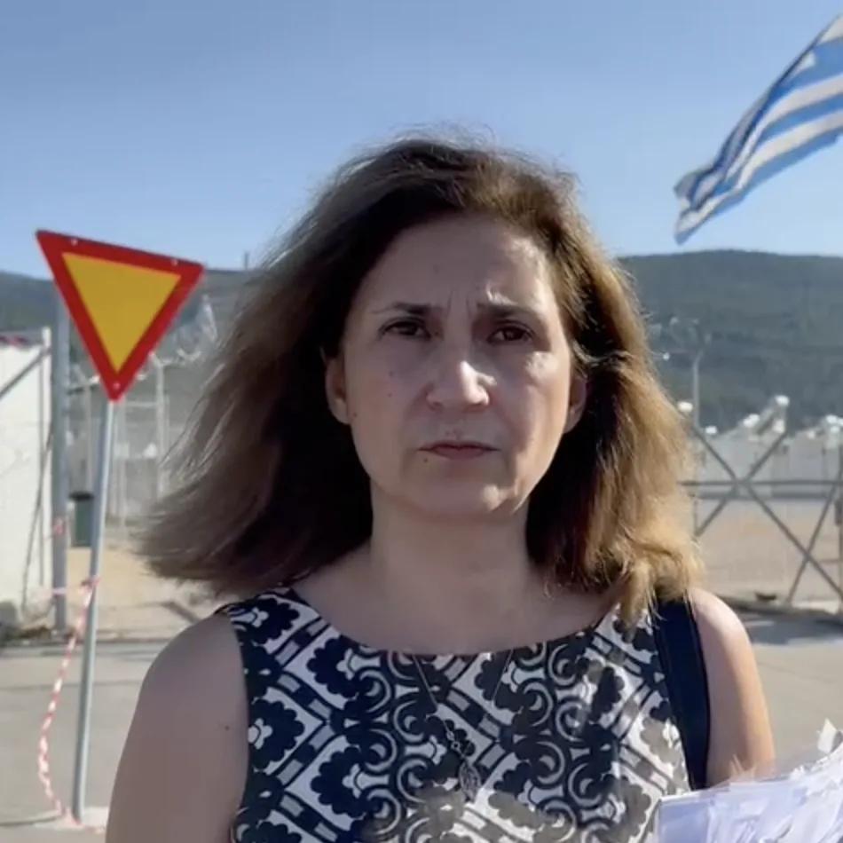 A person stands outdoors near a temporary settlement facility, with fencing, signage, and a blue-and-white flag visible in the background.
