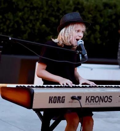 Max Keys wearing a black hat seated at a Korg Kronos keyboard, singing into a microphone during an outdoor musical performance.