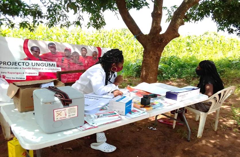 Community outreach worker seated at a table under a tree speaking with a young person, with supplies and informational materials arranged on the table.