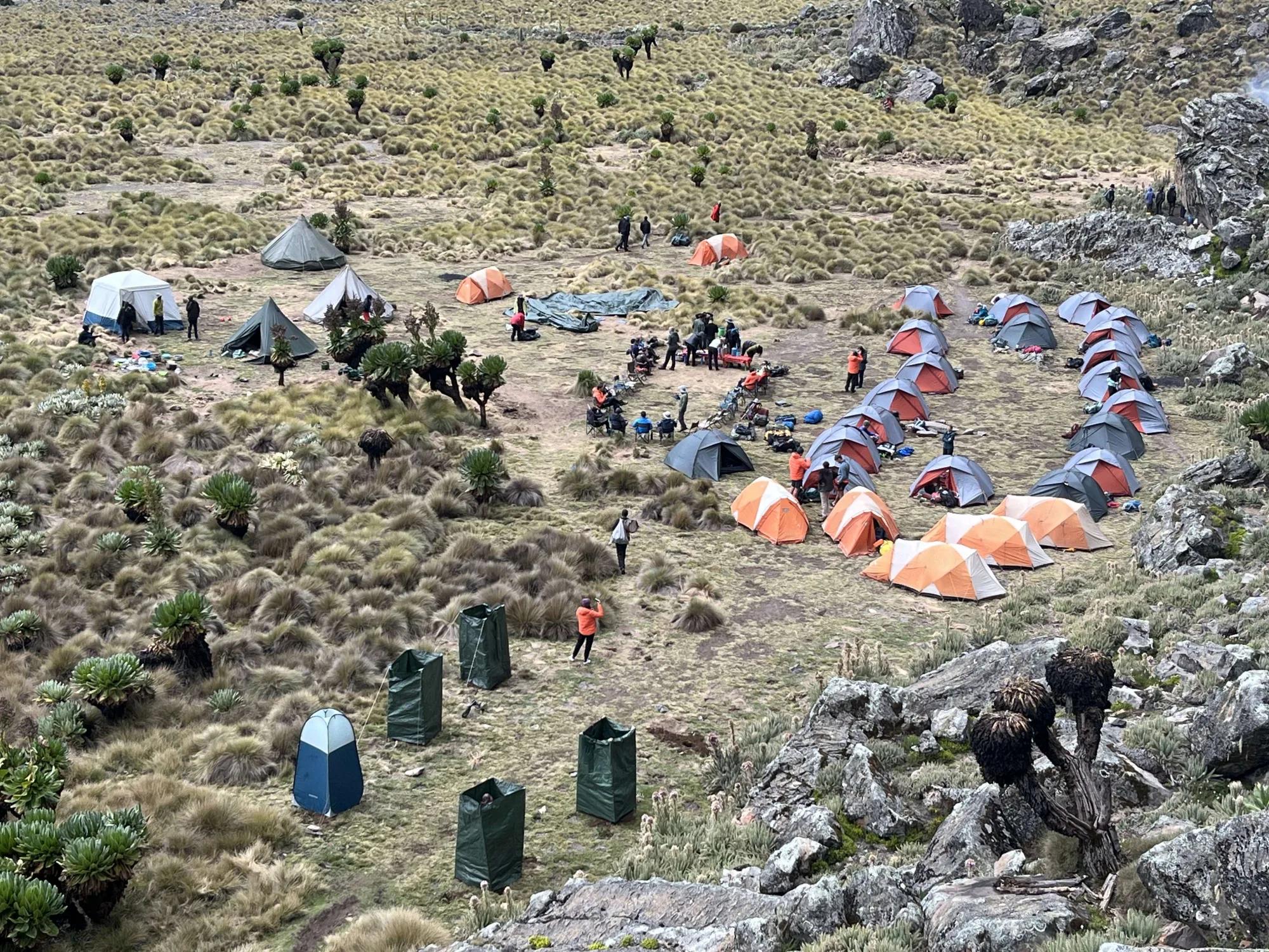 A group of grey and orange tents are clustered among scrub plants in a valley.