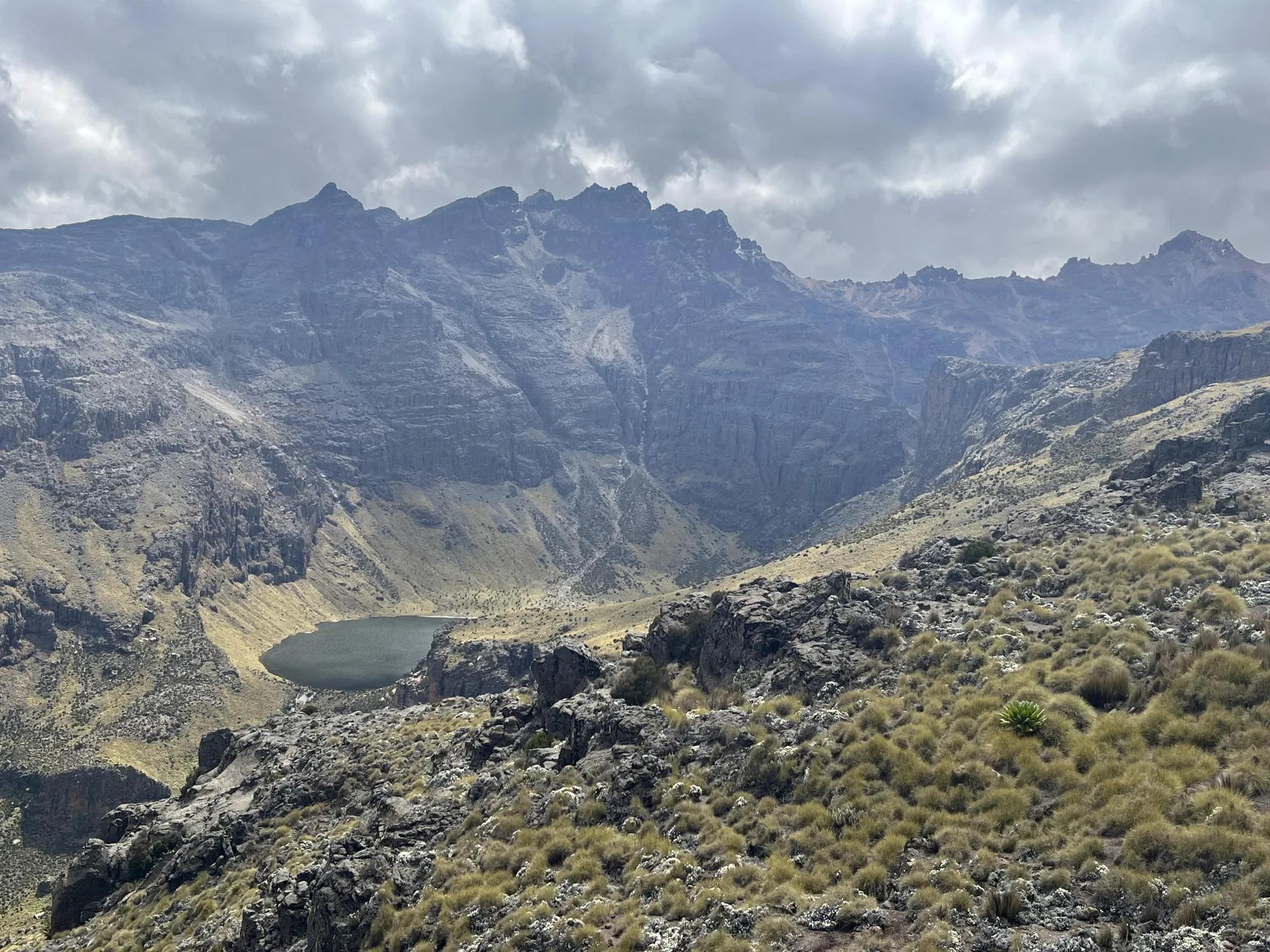 The photo shows a large mountain range against a cloudy sky, with a green valley and lake below, before the ground rises up to a corresponding mountain range at the bottom of the photo.