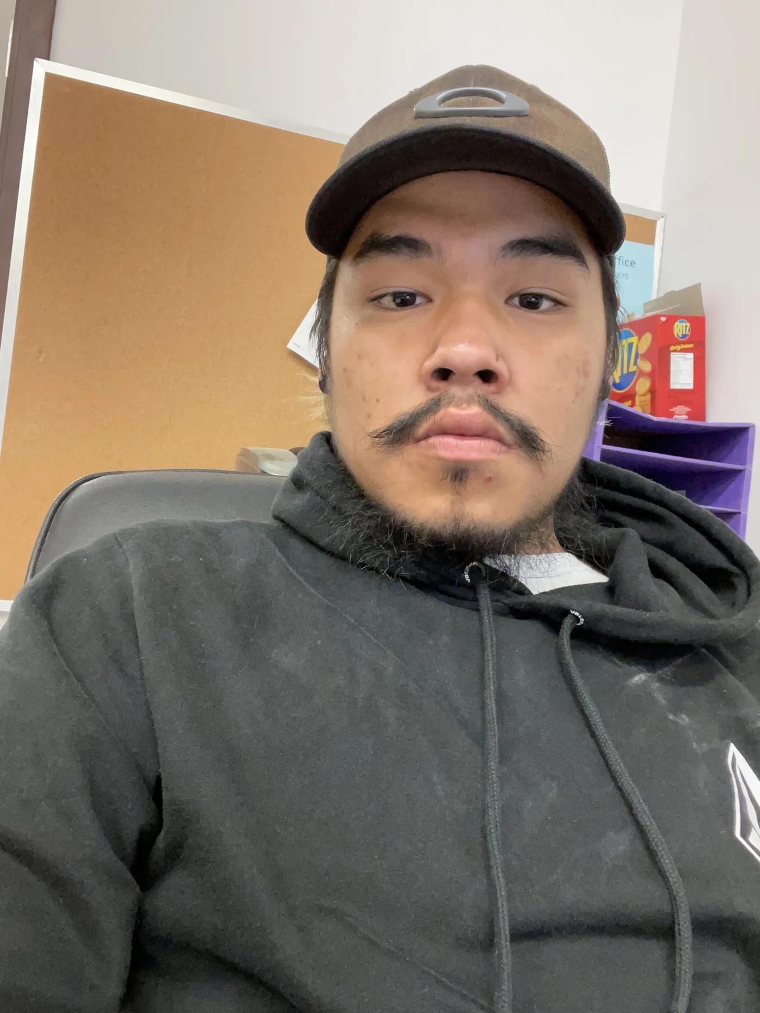 Close-up portrait of a person wearing a baseball cap and black hoodie, seated in an office setting with a bulletin board and storage shelves visible in the background.