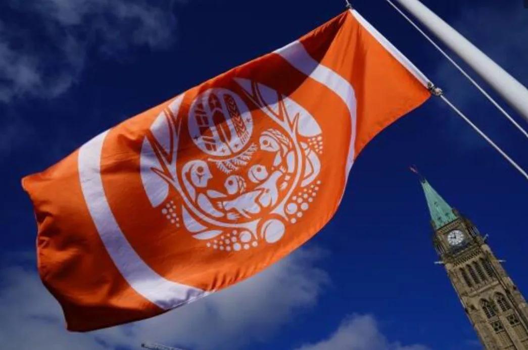 Orange commemorative flag featuring Indigenous-inspired artwork flying on a flagpole against a blue sky, with the Peace Tower of Parliament visible in the background.