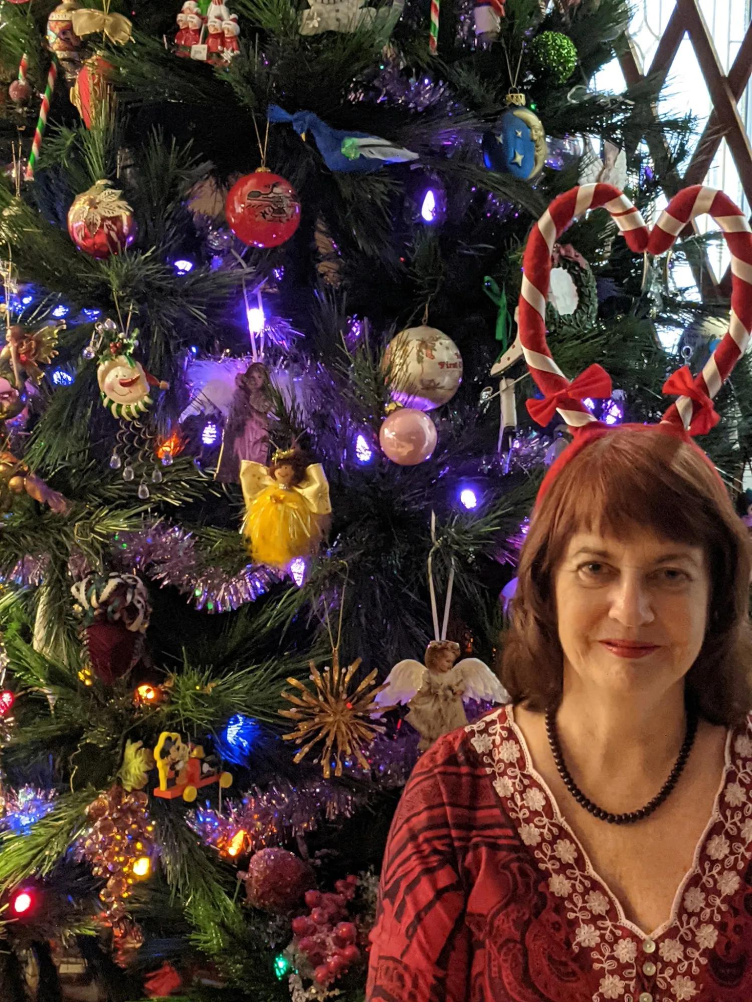 A person wearing a festive candy-cane headband stands in front of a Christmas tree decorated with colorful lights, ornaments, tinsel, and holiday figures.