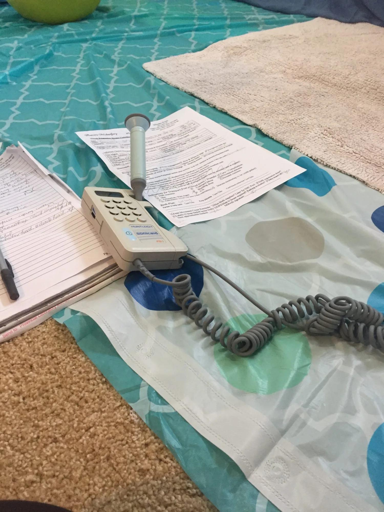 A portable fetal doppler device, coiled cord, medical forms, and a notebook are arranged on a bed with a patterned bedspread.