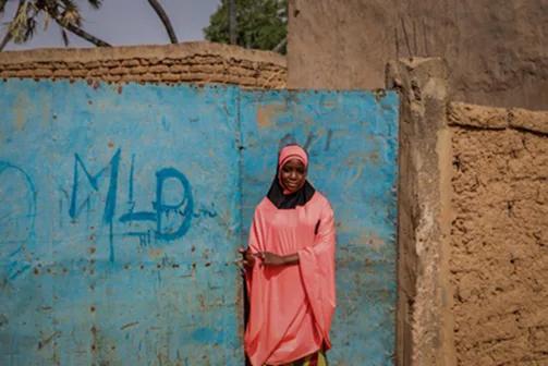 Individual wearing pink clothing and a head covering standing in front of a large blue gate set within an earthen wall in an outdoor community setting.