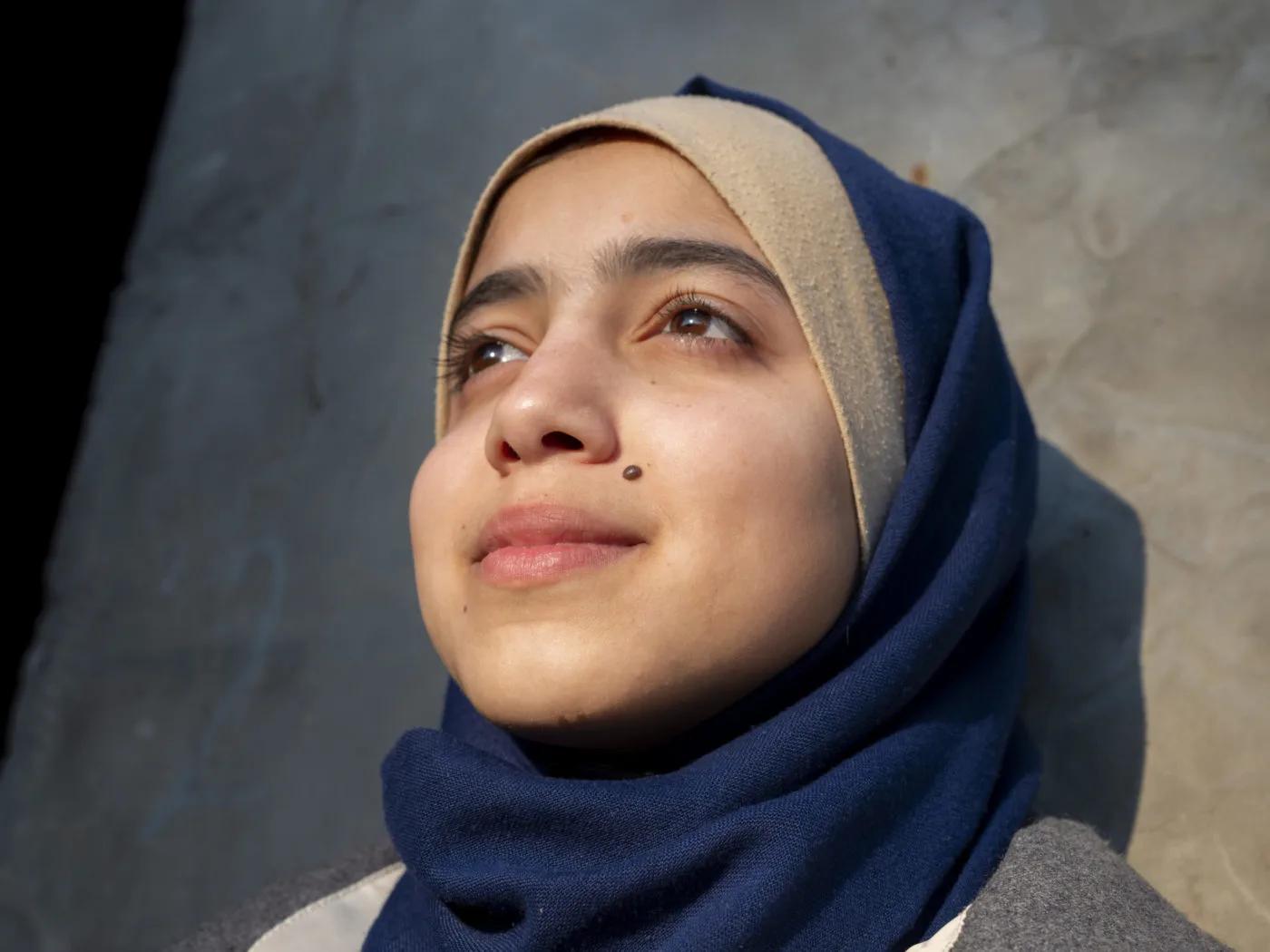 Close-up portrait of a student wearing a headscarf, standing outdoors and looking upward, with sunlight illuminating part of the face and a wall in the background.
