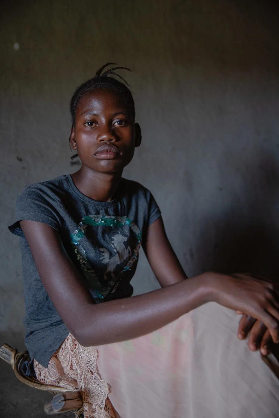 A seated student poses for a portrait in a dimly lit indoor setting, wearing a gray shirt and a light-colored skirt against a plain wall background.