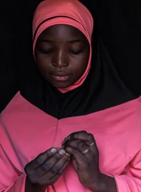Close-up portrait of an individual wearing pink and black clothing and head covering, looking down at their hands against a dark background.