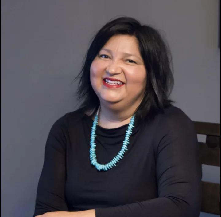 Studio-style portrait of a person seated indoors, wearing a black top and turquoise beaded necklace against a neutral background.