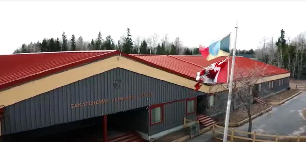 Exterior view of a school building with Canadian and community flags displayed in front of the entrance and trees in the background.
