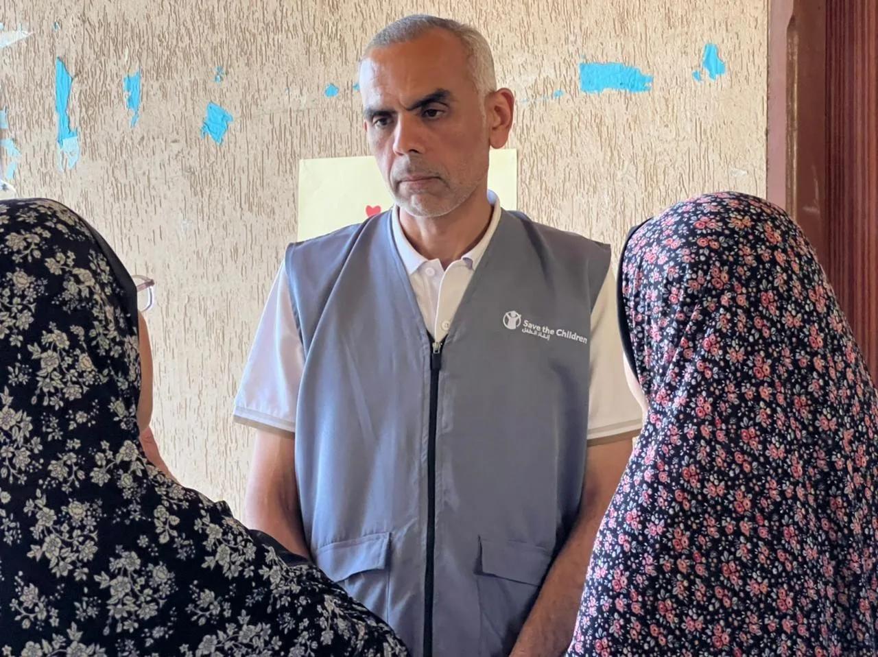 A Save the Children Representative Dr. Jamal Imam wearing a grey vest speaks with community members indoors while standing in front of a textured wall.