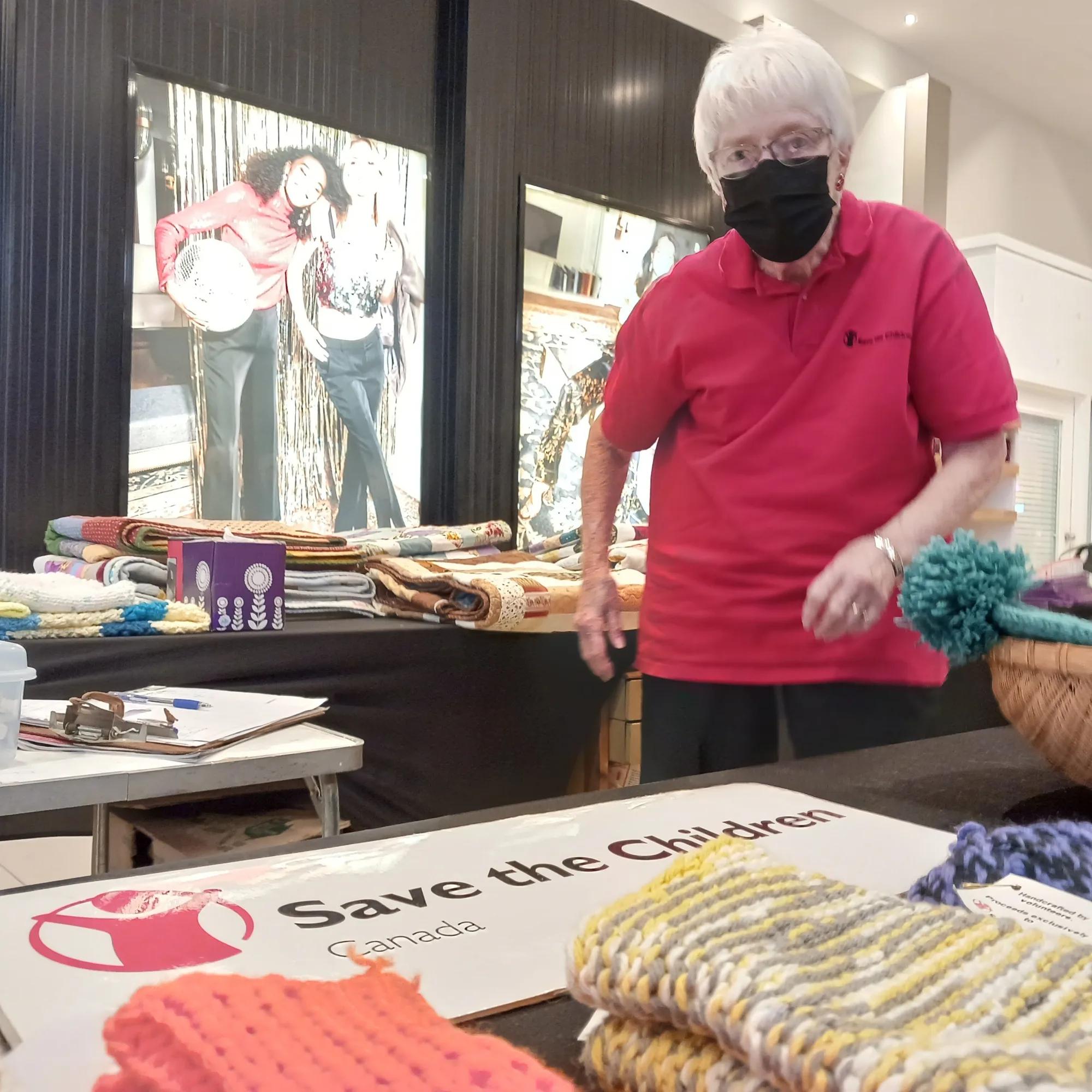 Person wearing a red Save the Children shirt and face mask standing behind a display table with knitted items, blankets, and a Save the Children Canada sign at an indoor fundraising event.