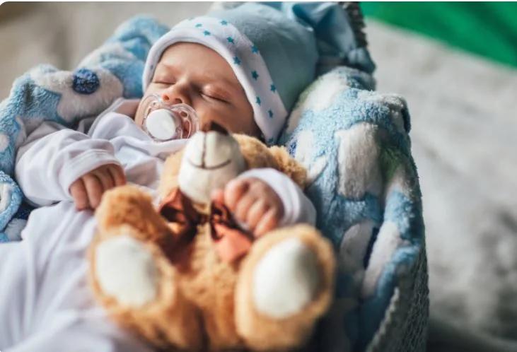 Close-up of a sleeping infant wrapped in a soft blue blanket, wearing a knit hat and holding a plush teddy bear while resting with a pacifier.