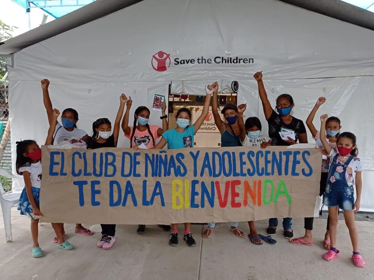 Group of children and youth standing in front of a Save the Children tent holding a large banner that reads ���El Club de Ni?�as y Adolescentes Te Da La Bienvenida,��� with several participants raising their hands and wearing face masks.