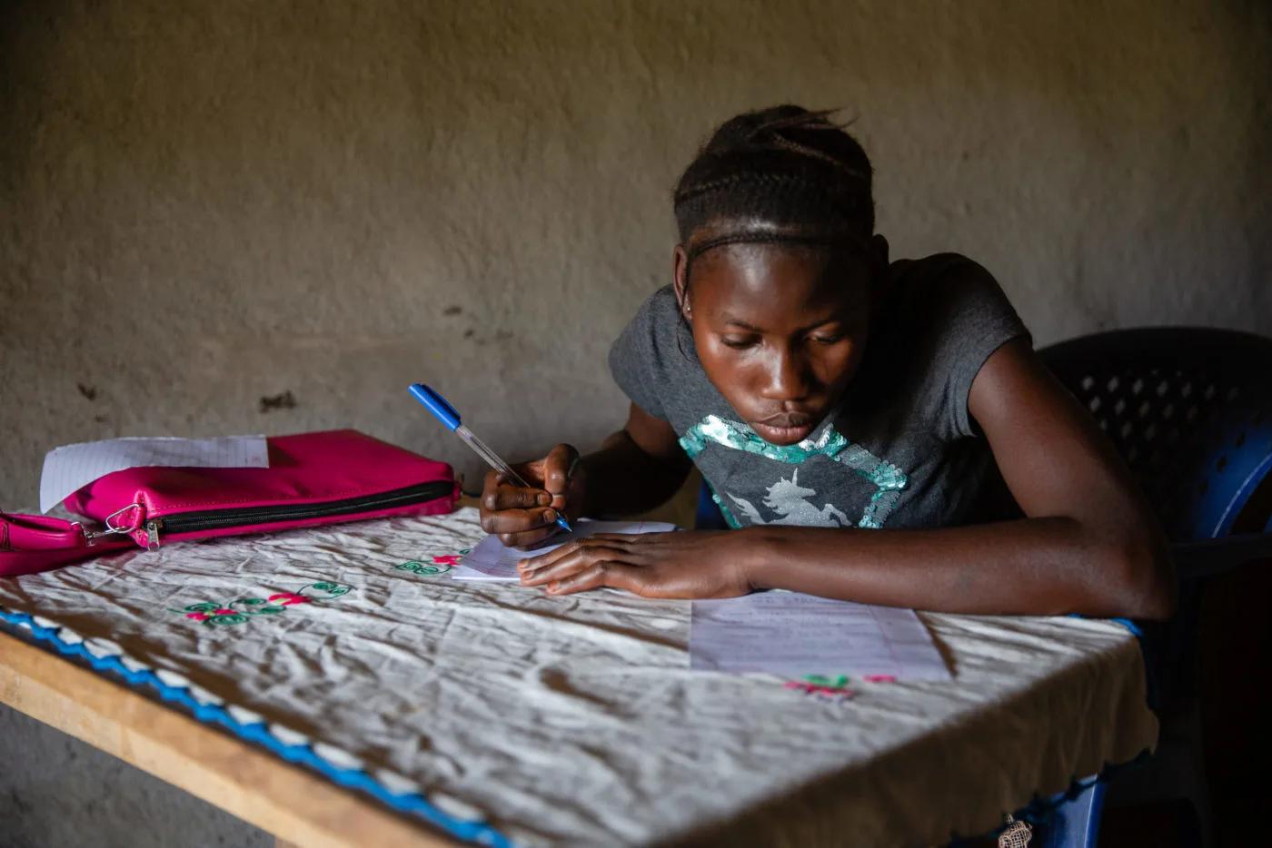 A student sits at a table writing on sheets of paper with a pen. A notebook and a pink pencil case are on the table in a modest indoor setting.