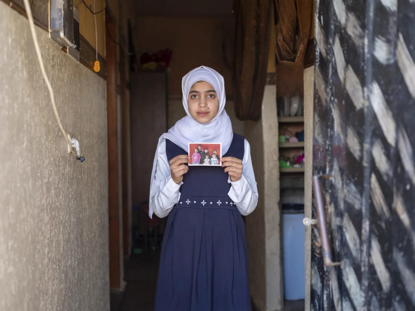 A child wearing a school uniform stands in a doorway holding a small family photograph, with the interior of a home visible in the background.