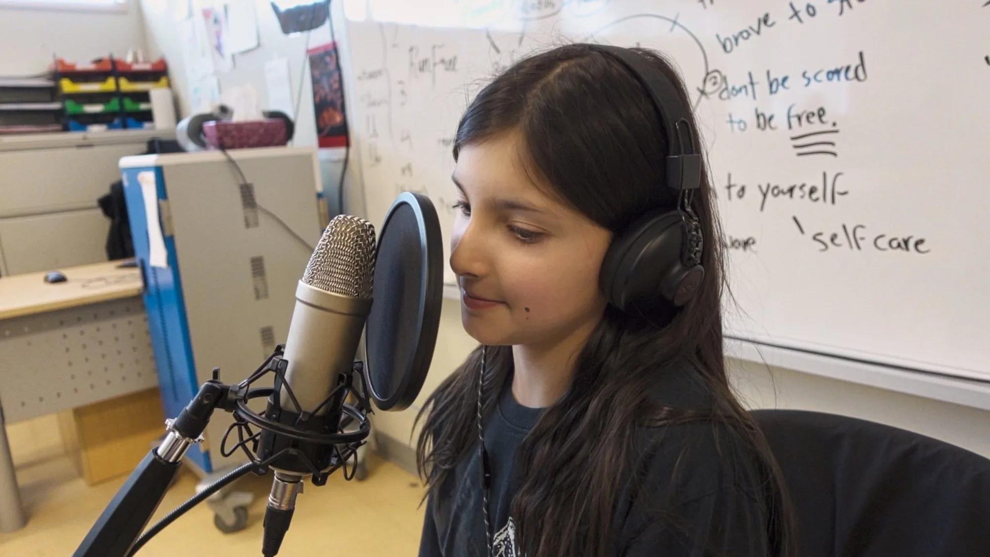 A young person wearing headphones speaks into a microphone with a pop filter while seated in a classroom, with a whiteboard and educational materials visible in the background.