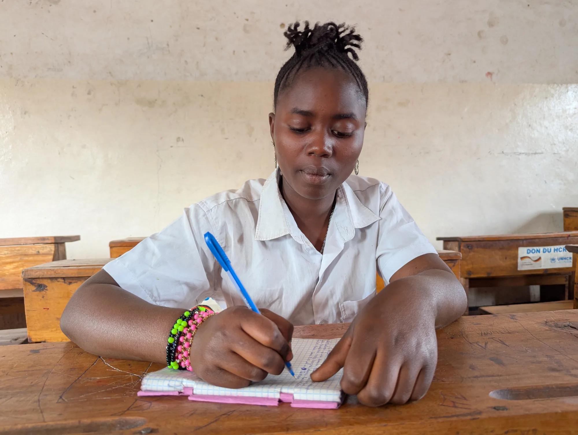 Student seated at a wooden desk writing in a notebook inside a classroom, with additional desks visible in the background.
