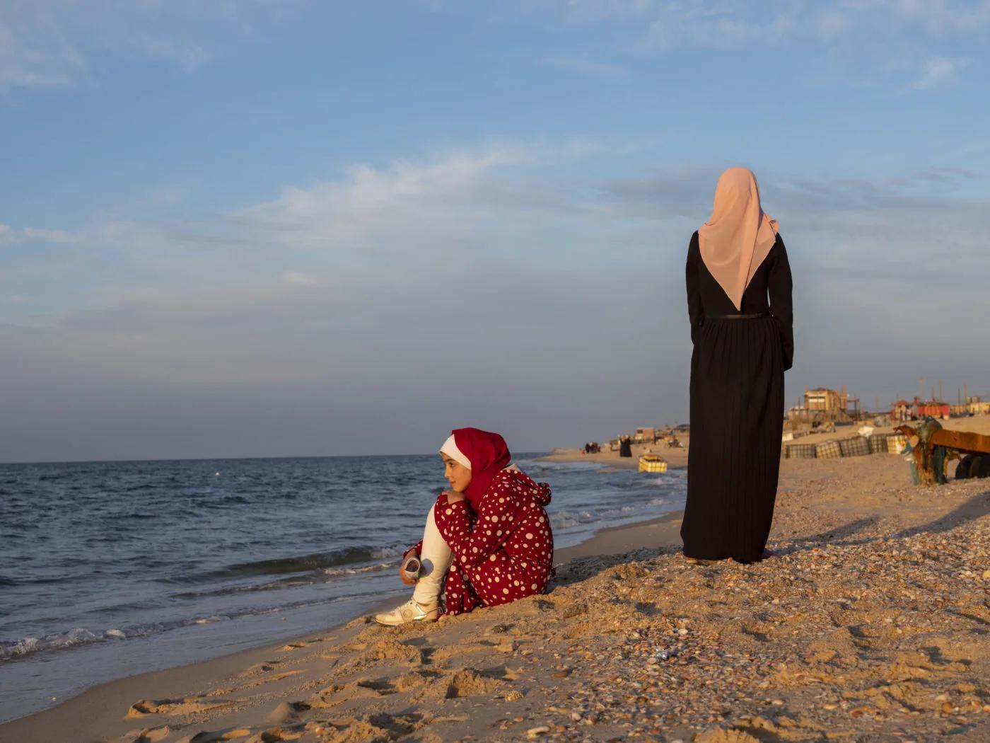 Two students on a sandy beach near the water's edge at sunset, with one seated facing the sea and another standing nearby. Buildings and other beachgoers are visible in the distance.