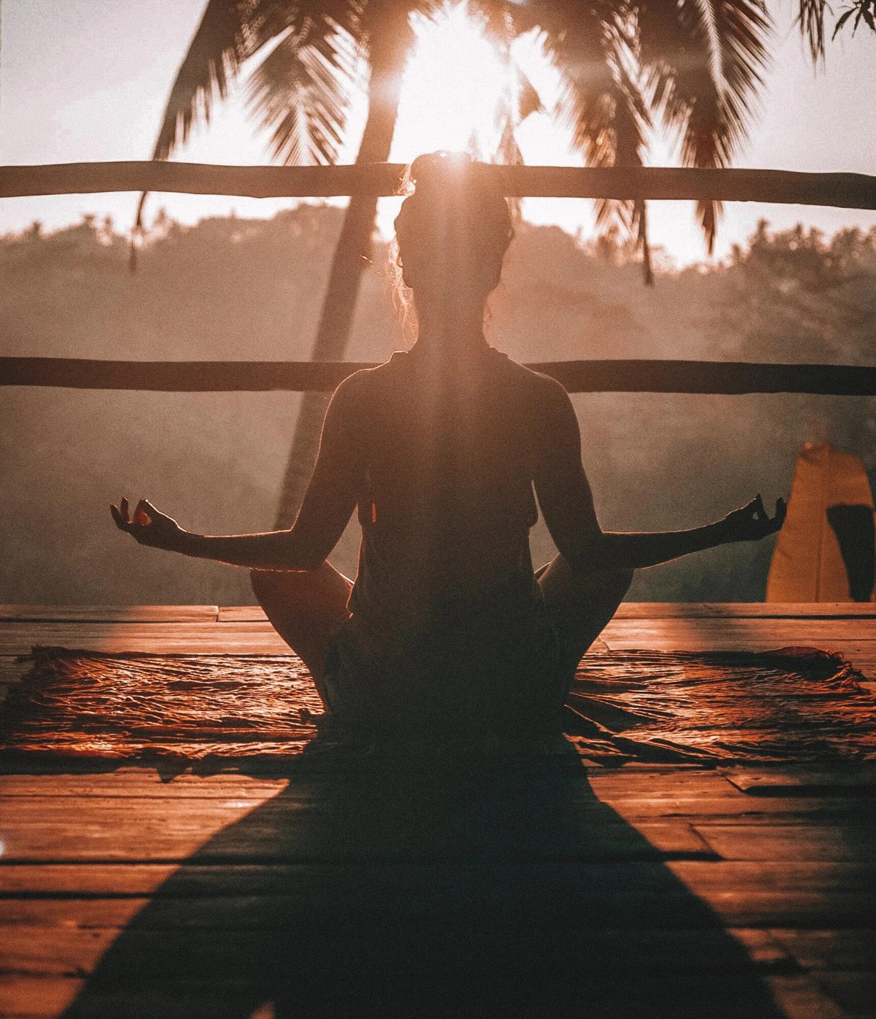 Silhouette of a person seated in a cross-legged meditation pose on a wooden deck at sunrise, with palm trees and warm sunlight illuminating the scene.