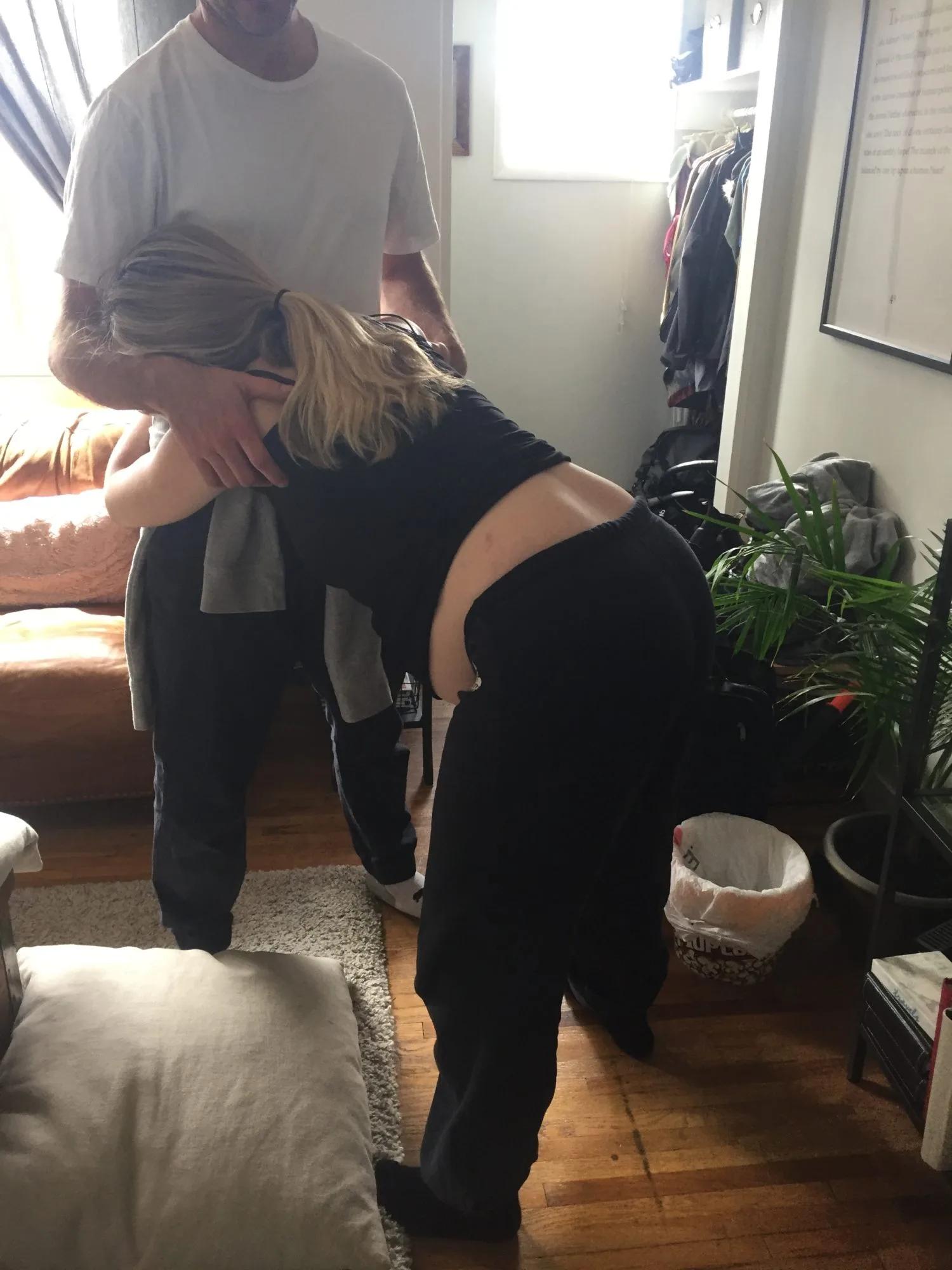 A birthing person leans forward while supported by another adult standing behind in a home setting, demonstrating a physical support technique commonly used during labor.