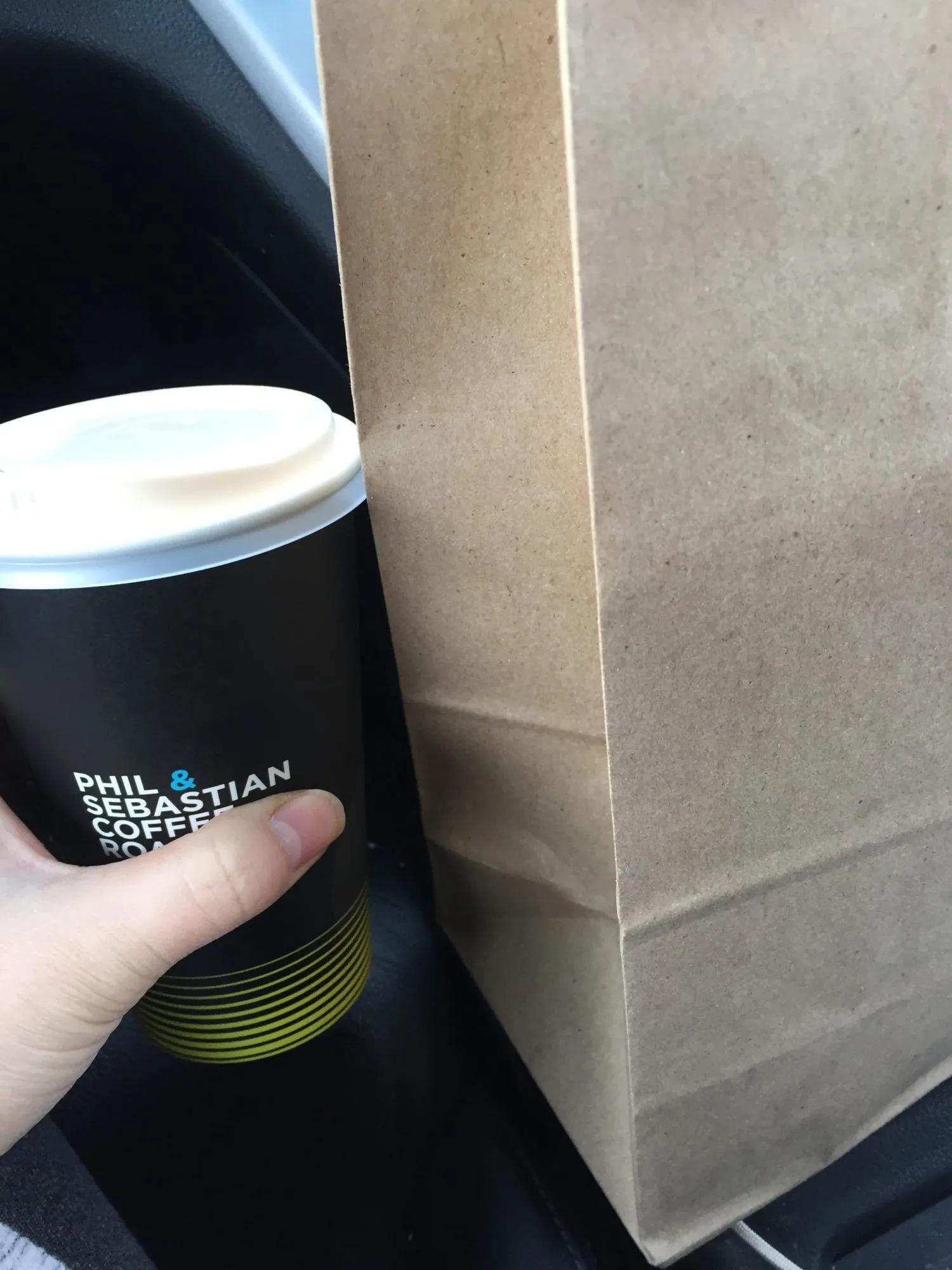 A hand holds a takeaway coffee cup beside a brown paper bag resting on a vehicle seat.