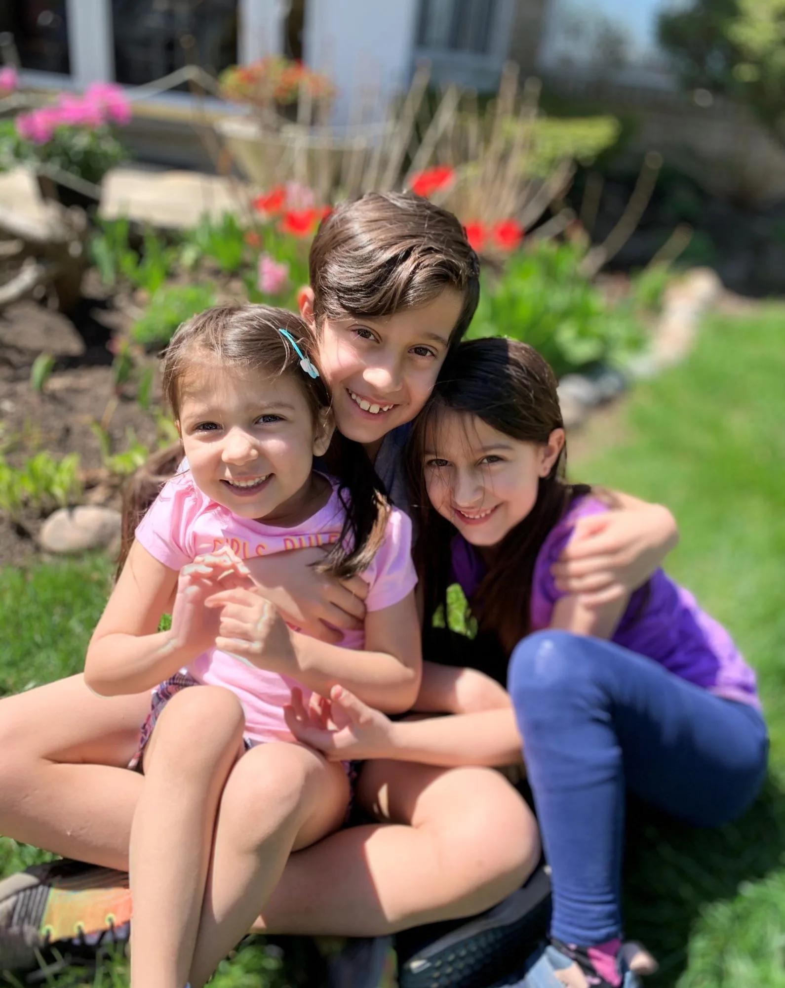 Three children sit closely together on a lawn in a residential garden, posing for a photo with flowers and greenery in the background.